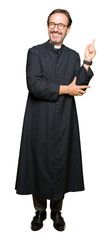 Middle age priest man wearing catholic robe with a big smile on face, pointing with hand and finger...