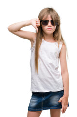 Young blonde toddler wearing sunglasses with angry face, negative sign showing dislike with thumbs down, rejection concept