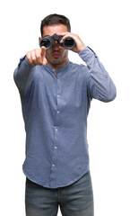 Handsome young man looking through binoculars pointing with finger to the camera and to you, hand...