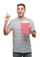 Handsome young man eating popcorn surprised with an idea or question pointing finger with happy...