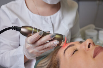A cosmetologist in rubber gloves does the rf-lifting procedure on the face. In the background, the...