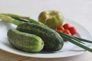 White Plate with fresh vegetables on wooden table. Two cucumbers, red cherry tomatoes, green cracked tomato with vertical splits, green Chives.  Healthy food, side view, copy space