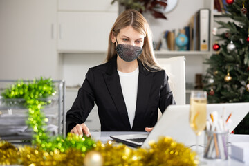 Woman employee in face mask working in office with documents and laptop during Christmas time