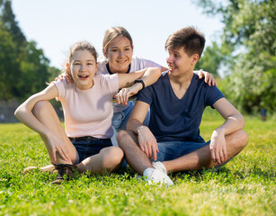 Fototapeta premium Cheerful teen friends gaily spending time outdoors on sunny summer day, sitting together on green lawn
