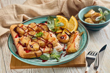 Tasty waffles with caramelized banana, mint, jam and sweet sauce on plate on wooden background.  Sweet meal. Dessert