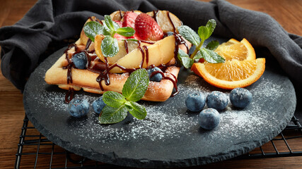 Tasty waffles with caramelized pear, berries, sweet sauce on black serving board on dark brown background. Dessert. Serving food