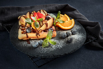 Tasty waffles with caramelized pear, berries, sweet sauce on black serving board on dark brown background. Dessert. Serving food