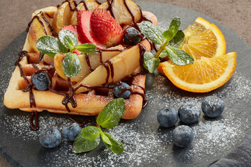 Tasty waffles with caramelized pear, berries, sweet sauce on black serving board on brown background. Dessert. Serving food