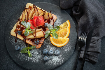 Tasty waffles with caramelized pear, berries, sweet sauce on black serving board on grey background. Dessert. Serving food