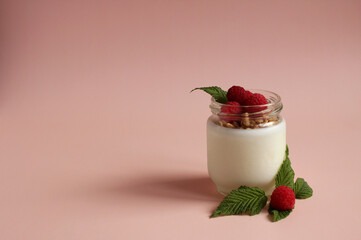 natural white yogurt in a glass jar with raspberries, muesli on a pink background