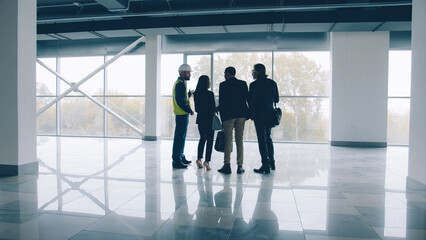 Group of engineers male and female are discussing new building interior checking blueprint and talking standing against panoramic window background