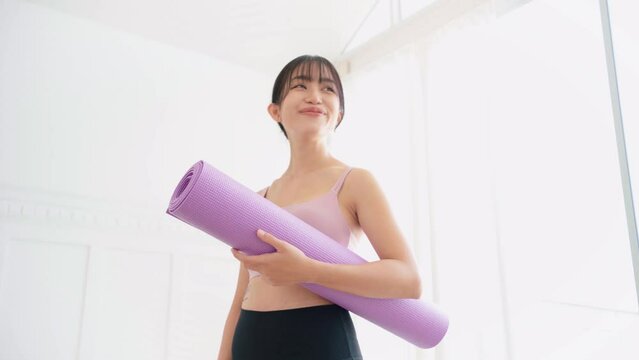 Smiling Asian woman wearing sports bra and yoga pants holding a rolled-up yoga mat standing in a bedroom with sunlight from the window. Real time portrait video.