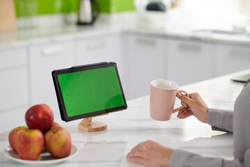 Green screen of tablet standing on wooden holder on white marble kitchen table