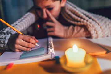 Little boy studying in low light with a burning candle.  Power outage, energy crisis concept.