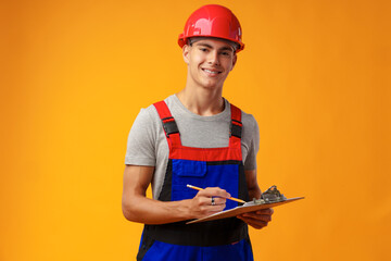 Fototapeta premium Young construction worker wearing hardhat and holding a clipboard on yellow background in studio