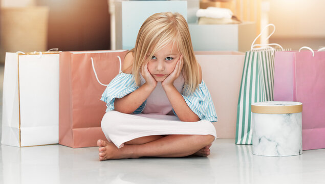 Bored child, fashion and shopping bags waiting on floor in kids clothing shop, store or boutique with cute girl sitting looking upset. Portrait of kid shopper or customer with sale purchase packages