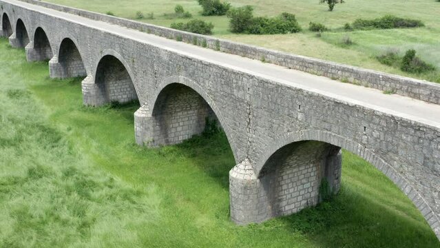 Old arch bridge made of stone over a green grassy field. Long vintage viaduct across a meadow in the valley, where once was a lake or wetland. Tsar's bridge (Carev most) near Niksic in Montenegro.