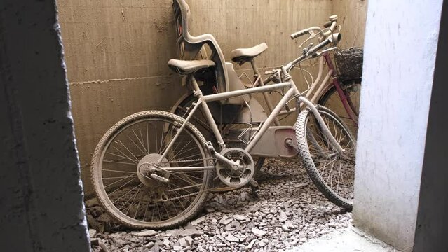 Abandoned bikes with flat tires left leaning on a sandy wall, on rubble covered ground. With a child seat, covered in dirt