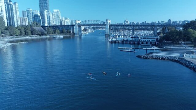 Kayak outrigger float on left side birds top view aerial flyover 5 people at foot of English Bay False Creek in Vancouver BC as they huddle together before the paddle away on clear fall afternoon