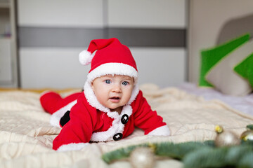 Cute Caucasian baby girl 3-4 months old in Santa costume lying on knitted cozy blanket on bed with decorated fir tree. Merry Christmas xmas and happy new year 2023