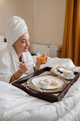 Pretty caucasian female in bathrobe and towel on head having a breakfast in bed in the morning at the hotel room - 536251692
