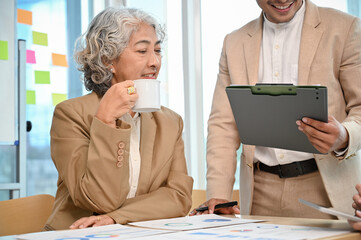 Professional aged Asian female boss giving advice to her male staff while sipping her coffee