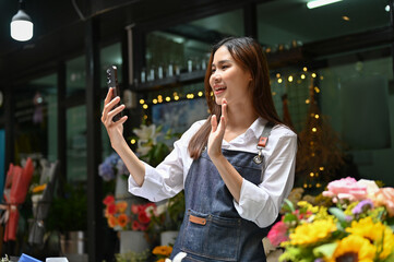 Charming Asian female florist using smartphone, streaming live on her flower shop's social media.