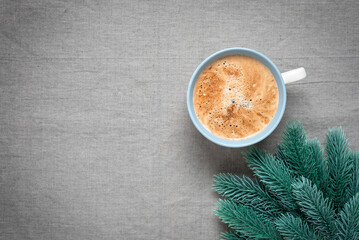Cup of cappuccino coffee and decorative spruce branch, gray linen texture background. Cozy Christmas background. Top view, selective focus.