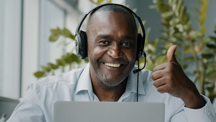 Senior african employee sit at office desk wear headphones with microphone connect to video conference business call on laptop communicate by webcam talk in online chat does thumb up approval gesture