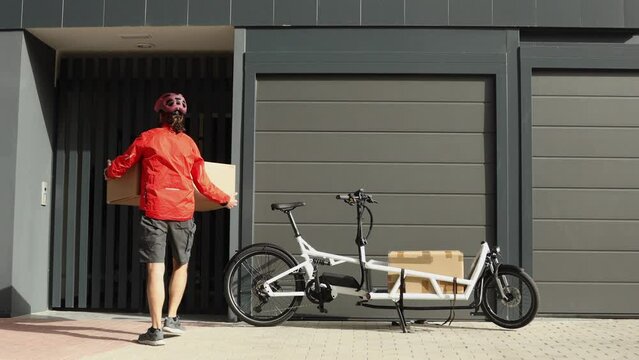 Young courier with red clothing and helmet riding cargo bike arriving at the shipping destination to deliver a package to a city address, european housing, horizontal video
