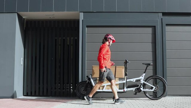 young courier with red clothes and helmet riding cargo bike arriving at the shipping destination to deliver a package to an address in the city, the recipient is not at home, horizontal video