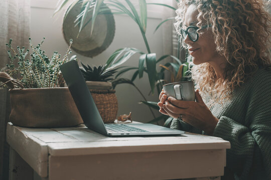 One Lady Using Laptop At Home Drinking Tea Or Coffee In Indoor Leisure Activity Online. Pretty Woman Working On Computer And Smiling. Lady Surfing The Web Alone With Nature Plants Garden In Background