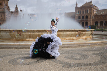 Young teenage woman in white suit with black polka dots, dancing flamenco in front of water fountain and surrounded by soap bubbles. Flamenco concept, dance, art, typical Spanish dance, bubbles. - obrazy, fototapety, plakaty