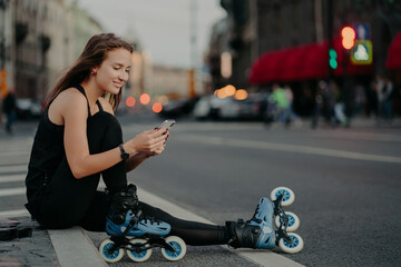 Pleased sporty woman wears sportsclothes rollerblades sits on road checks newsfeed via smartphone...