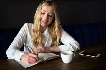 Attractive blonde girl in a white shirt - writes in a notebook. In a gray cafe on a blue sofa at a brown table.