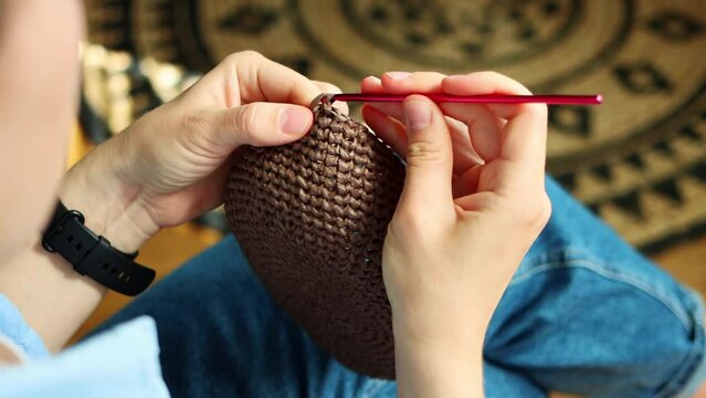 Cropped young woman crocheting brown bag using pink hook and raffia yarn sitting at cozy home. Earning money on your handmade hobby concept. Knitting is tranquil method of calming the nervous system