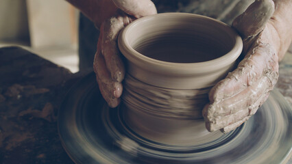 Senior male ceramist is concentrated on making pot from clay on spinning throwing wheel. He is...