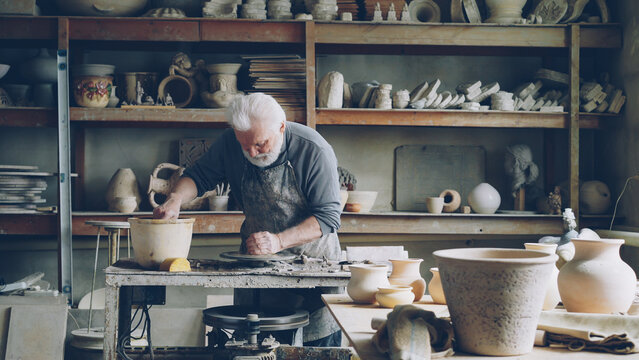 Hardworking silver-haired man is working with clay on potter's wheel in workplace, shaping piece of loam. Beautiful ceramic utensils, handmade pots and vases on shelves are visible.