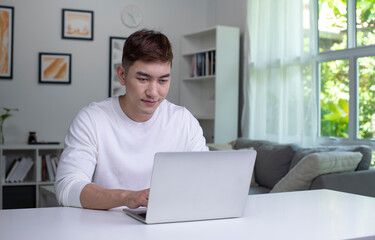 Young Asian man using laptop at home, looking at screen, chatting, reading or writing email, sitting on desk, serious male student doing homework, working on research project online.