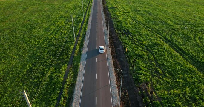 Aerial view of a white car moving along an asphalt road. The drone follows the car in the countryside. Paved road with markings. Highway in a green field. Sunlight at sunset.