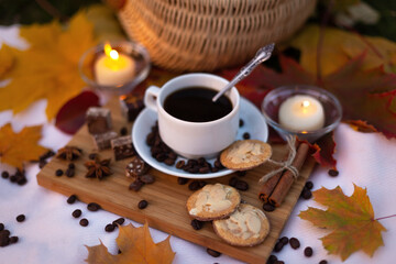 Cup coffee with cookies and chocolate on background of burning candles in evening autumn park with maple leaves. Still life. Picnic outdoors
