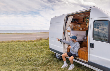 Hombre viajero caucásico, sentado en la camper, buscando ubicación en su teléfono móvil y señalando al paisaje.