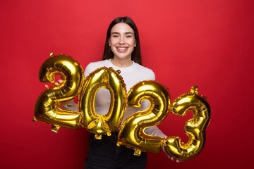 A young woman with a wide smile holds the numbers 2023 from balloons on a red background. New...