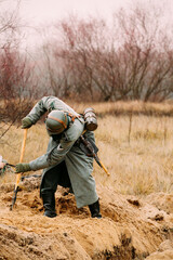 Re-enactor Dressed As German Wehrmacht Infantry Soldier In World War Ii With Shovel Digs Trench. Arrangement Of A Defensive Position. German Military Dress Of A German Soldier At World War II.