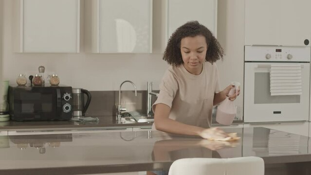 Medium slowmo of young Biracial woman using sprayer and wiping cloth while cleaning kitchen table, doing household chores in apartment