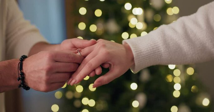 Close-up of guy proposing to girl. He holds the girl's hand and puts wedding ring on finger. The girl has long nails and nude manicure. Christmas tree flickers behind. Engagement.
