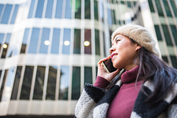 Fototapeta premium Side view of content Chinese female in outerwear and beret having phone conversation while standing near modern multistory building in city