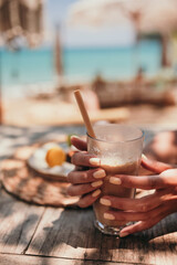 Woman hands with yellow manicure hold ice coffee latte in tall glass with straw by the sea in beach bar
