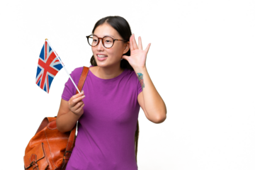 Young Asian woman holding an United Kingdom flag over isolated background listening to something by putting hand on the ear