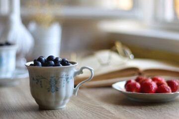 Vintage tea set filled with blueberries, blackberries and starwberries, open book, reading glasses and decorative candles on the table. Selective focus.